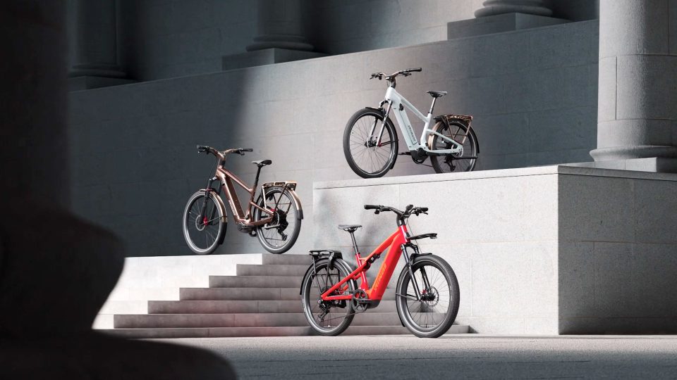 Three electric bicycles—including the Specialized Vado 3 in bronze, white, and red—are displayed on different levels of gray stone steps with columns in the background. Sunlight highlights the bikes and part of the stairs, capturing a sense of inertia and excitement around this latest launch. Frame from Stash Magazine article.