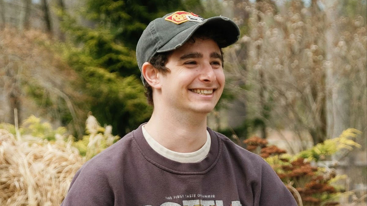 A young man wearing a dark baseball cap and purple sweatshirt smiles outdoors, with blurred trees and plants in the background—capturing Spencer Levin, Post Production Supervisor at Alkemy X, in a candid moment. Frame from Stash Magazine article.