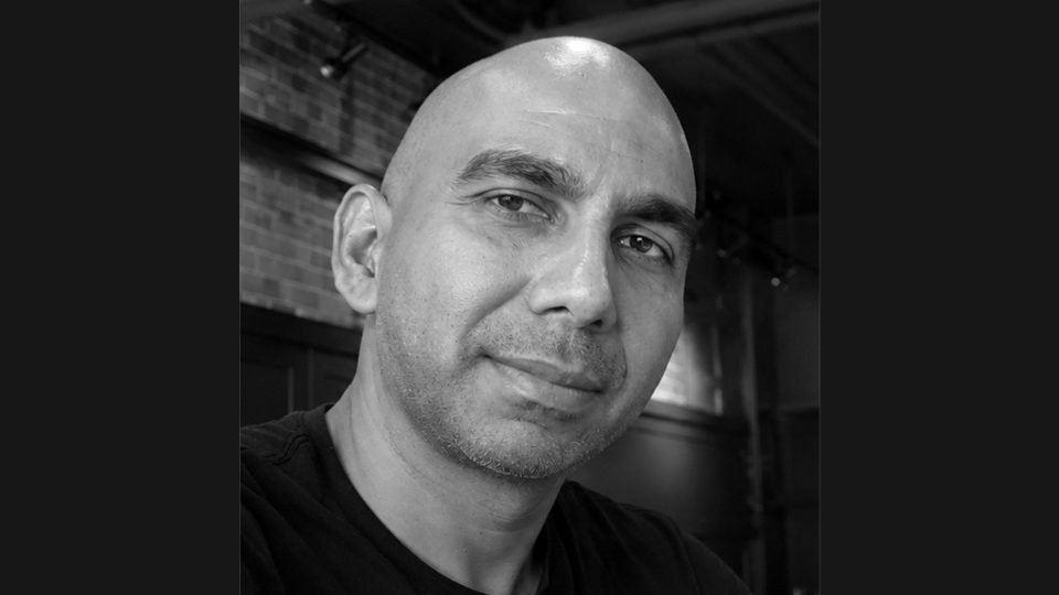 A bald man with a slight smile looks at the camera in this black-and-white portrait. Exposed brick and dark walls set the scene for Spiders-&-Sparrows-Signs-Emmy-Award-Winning-Director-Raj-Dias. Frame from Stash Magazine article.