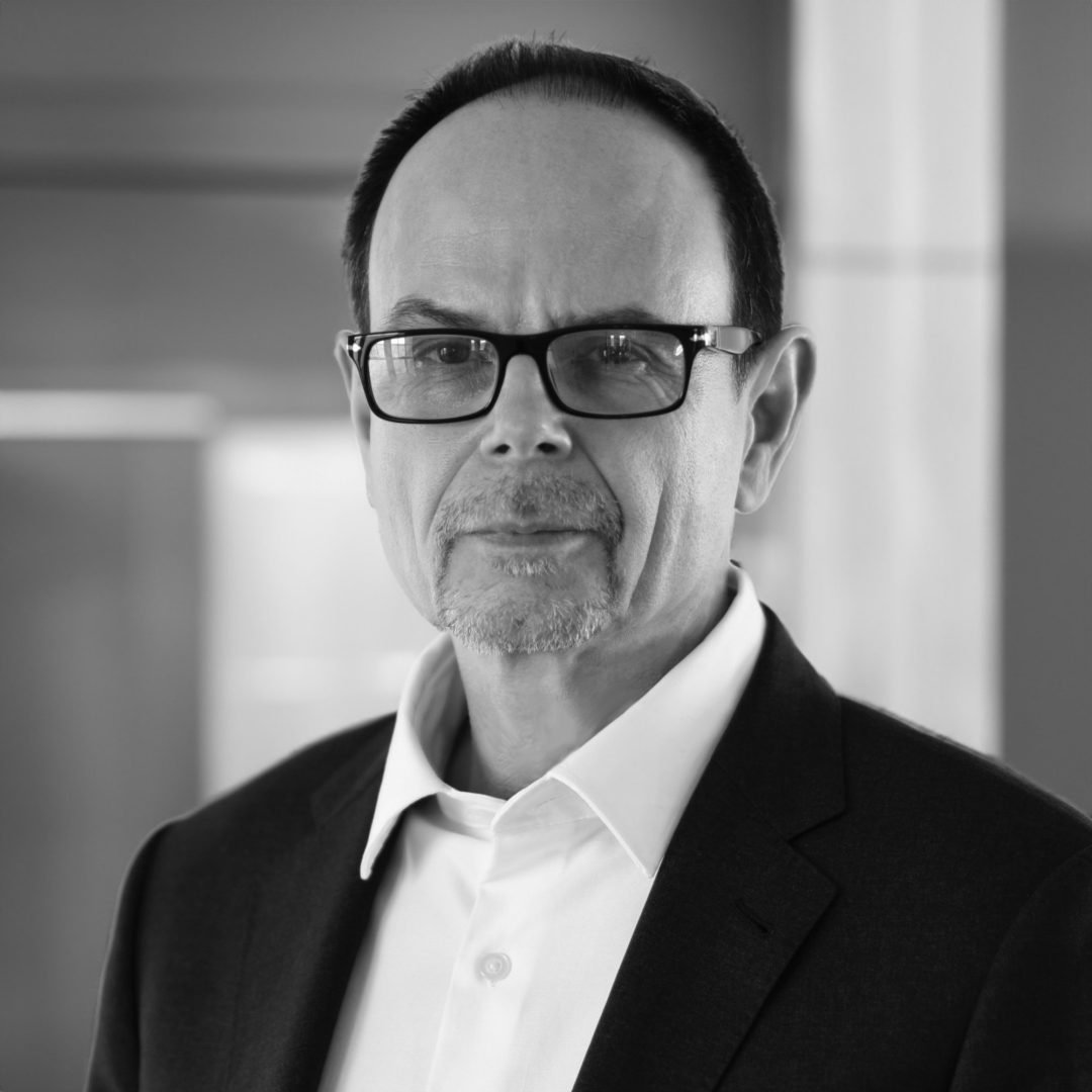 A middle-aged man with short dark hair, glasses, and a goatee wears a suit jacket over a white shirt, looking seriously at the camera in a modern indoor setting at OFFF 2026. Frame from Stash Magazine article.