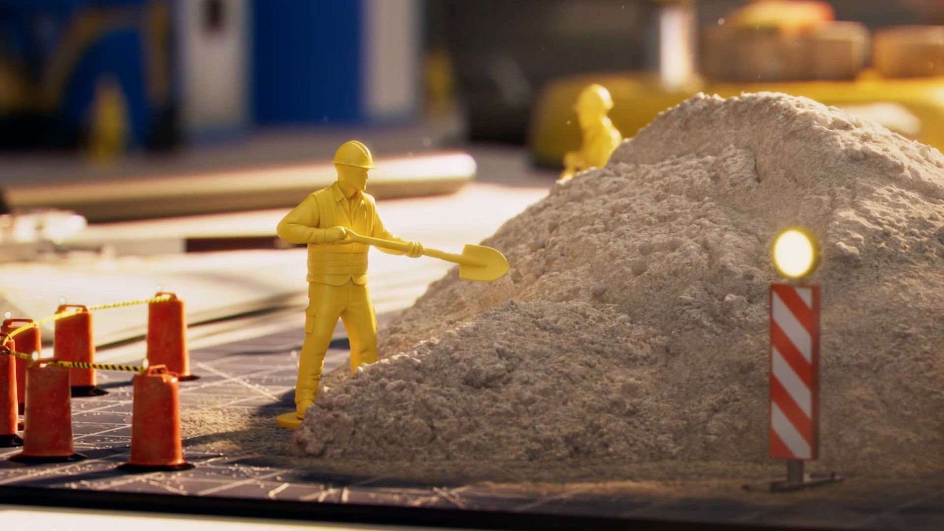 A yellow plastic construction worker figurine with a shovel stands next to a pile of sand, surrounded by orange barriers and an "Under Construction" sign from Studio Bump. Another figurine is visible in the background. Frame from Stash Magazine article.