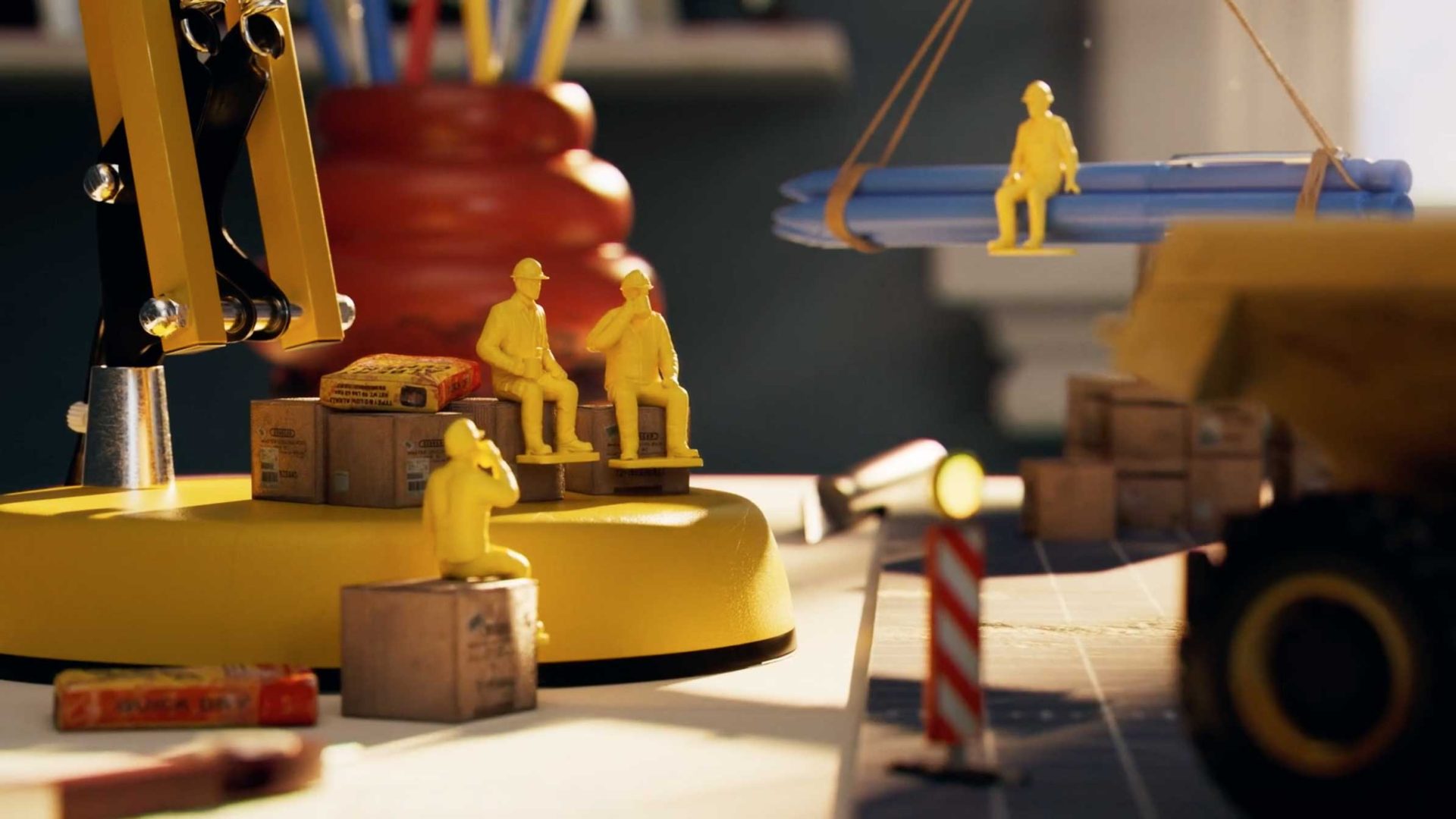 Miniature yellow construction worker figurines sit on boxes and are lifted by a small crane, creating an "Under Construction" vibe on a desk filled with office supplies. The playful scene is perfect for Studio Bump’s creative workspace. Frame from Stash Magazine article.