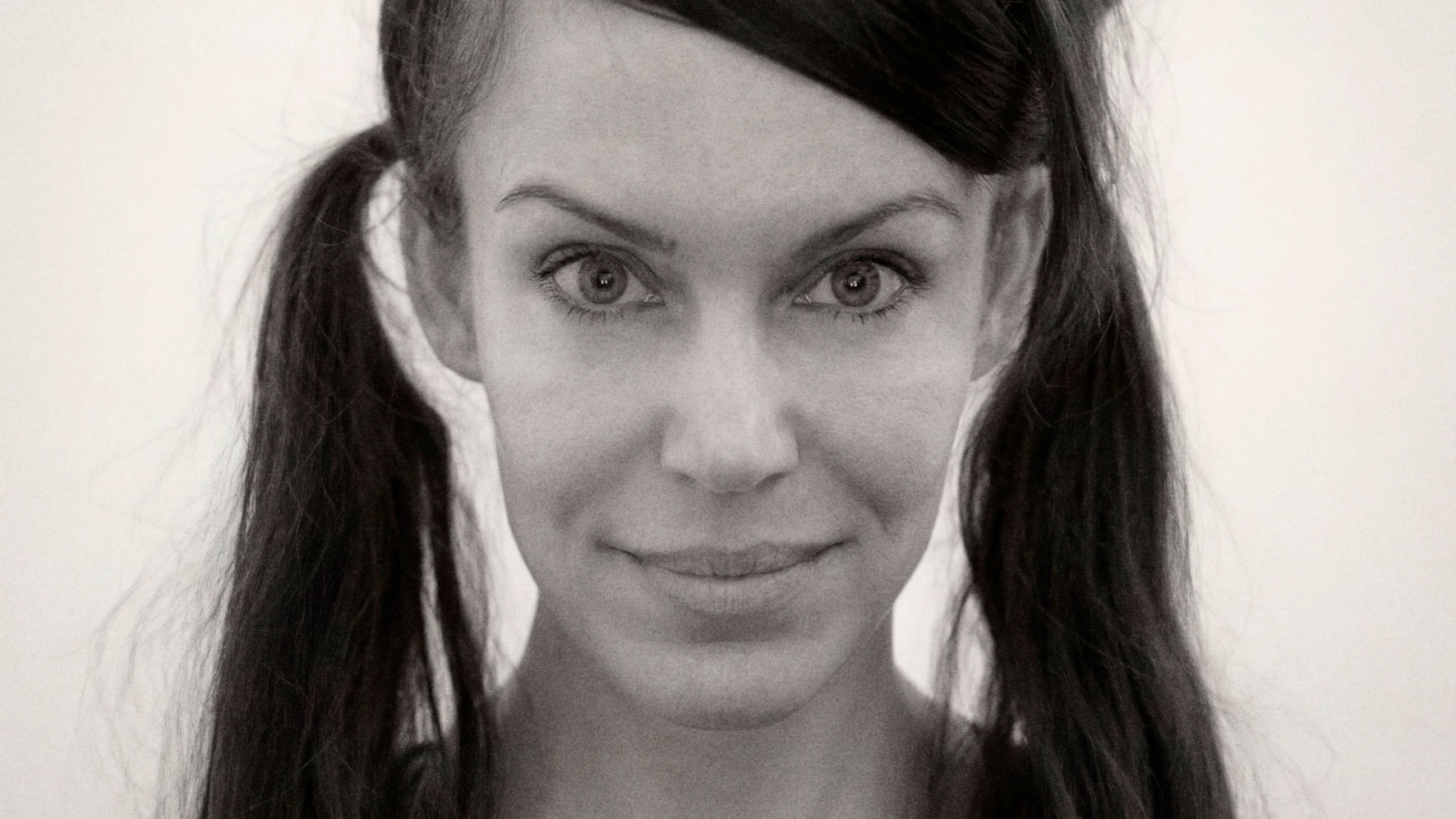 Black and white close-up of a woman with long dark hair in two pigtails, looking directly at the camera and smiling softly against a plain light background—capturing an elegant simplicity reminiscent of Studio Private’s signature style as they add Mikaela Westerholm as Head of Image. Frame from Stash Magazine article.