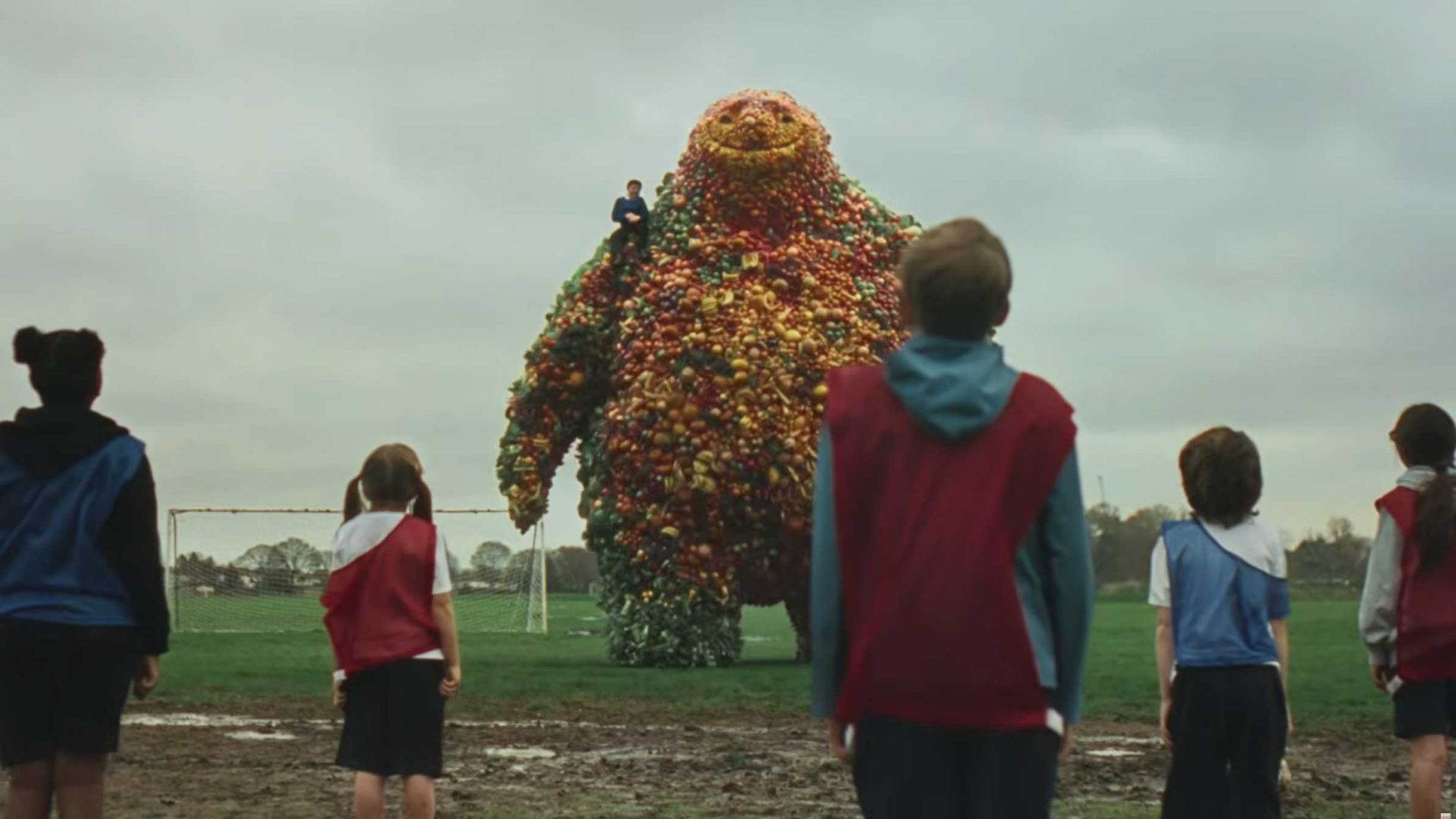 Several children stand on a muddy soccer field facing the Tesco Friendly Fruit Giant, a cheerful creature made of colorful balls, with someone perched on its shoulder. The overcast sky and distant soccer goals set the scene for feel-good food fun. Frame from Stash Magazine article.