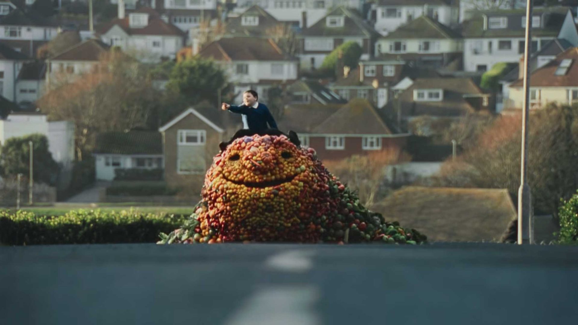 A person rides on top of a Friendly Fruit Giant made of apples, moving down a suburban street lined with houses—a true feel-good food adventure brought to life. Frame from Stash Magazine article.