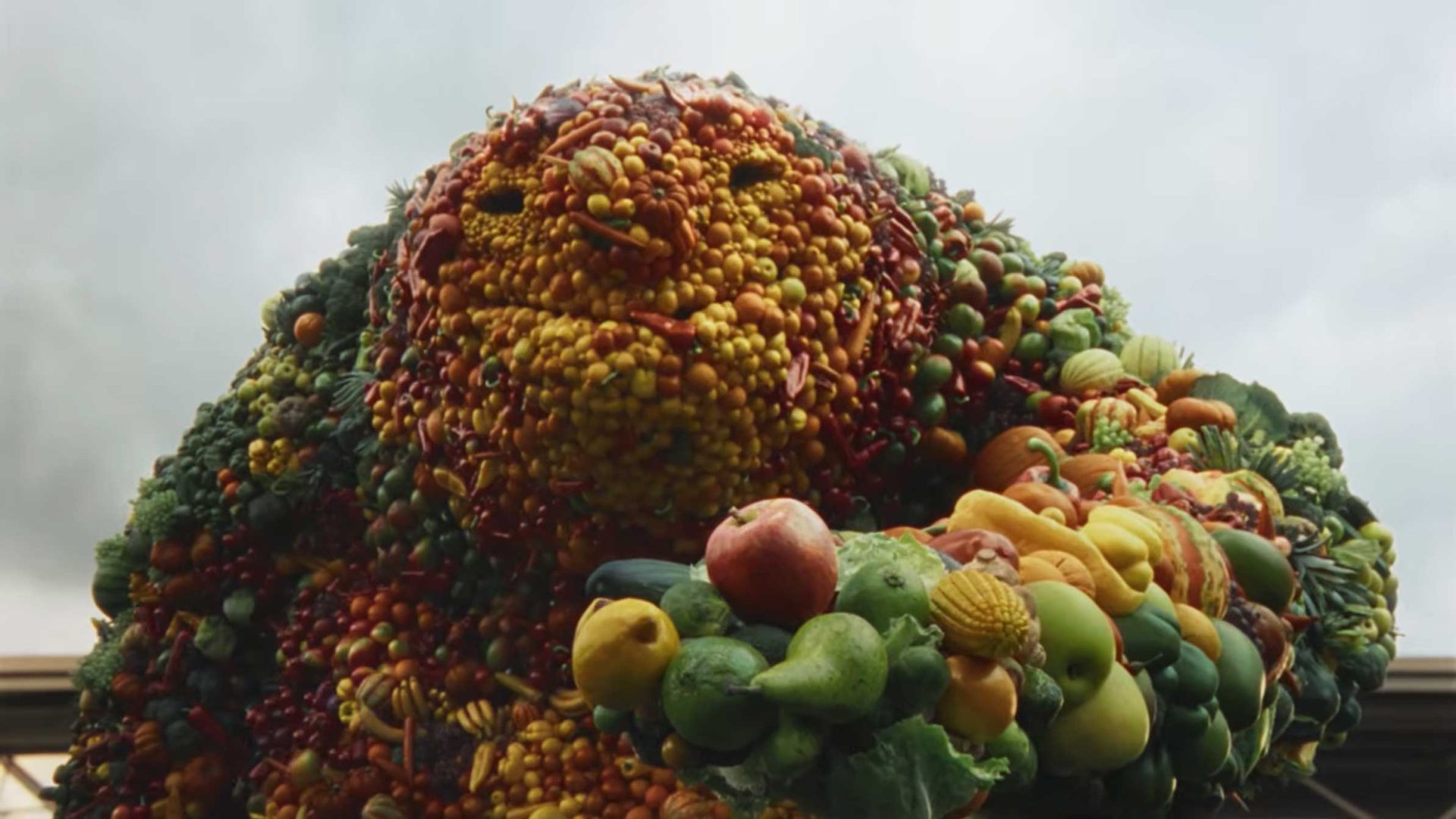 A large creature made entirely of various fruits and vegetables, including Tesco apples, peppers, and broccoli, stands outdoors against a cloudy sky. The produce forms a face and body, giving this Untold Studios creation a whimsical, feel-good food appearance. Frame from Stash Magazine article.