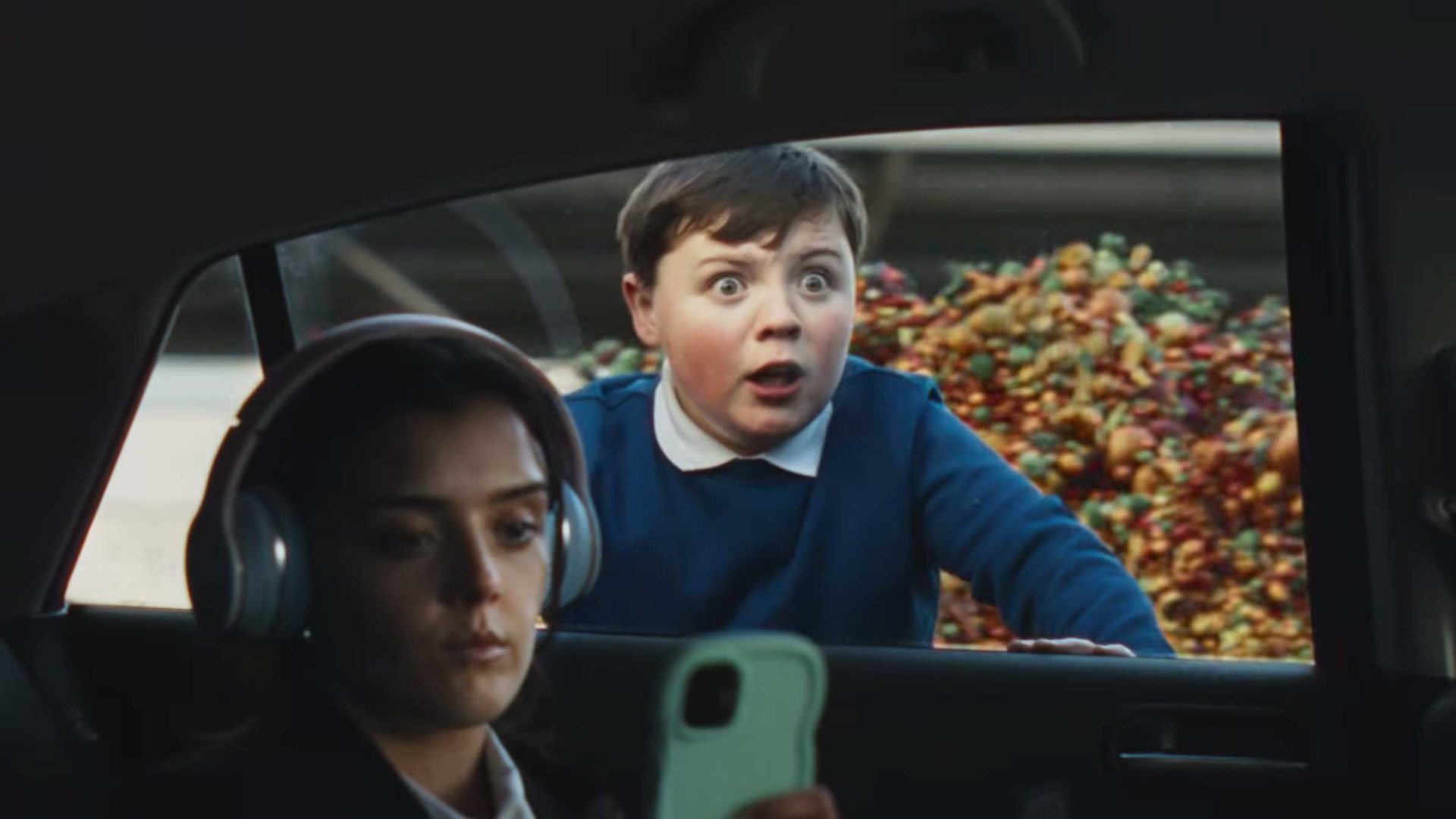 A girl wearing headphones looks at her phone inside a car, while a surprised boy outside—next to a pile of Tesco apples—peers through the open window with a shocked expression. Frame from Stash Magazine article.