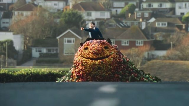 A person rides atop a Friendly Fruit Giant—Tesco’s feel-good food creation—smiling in the middle of a suburban road, surrounded by houses and lush greenery. Frame from Stash Magazine article.