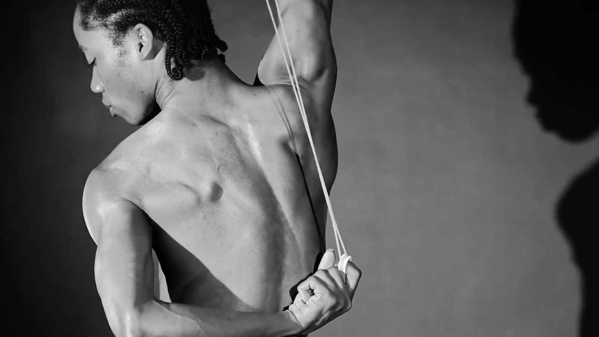 A person with braided hair stretches an elastic band behind their bare back. Their arm is raised, showing defined muscles and a shadow on the wall. The black and white image captures a moment of raw strength and artistry, reminiscent of the creative spirit found at University of the Arts London. Frame from Stash Magazine article.