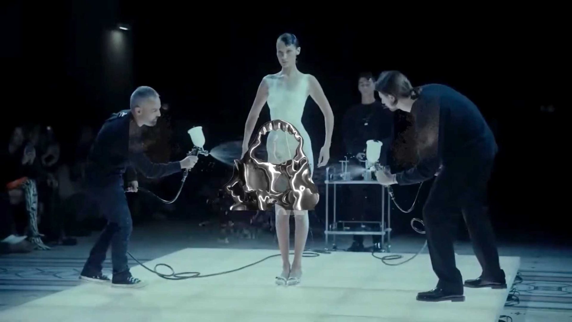 A woman stands on a platform as two people spray paint a shiny, metallic dress onto her body in a Midnight Club-inspired scene. The dark room, with onlookers in the background, exudes an avant-garde energy reminiscent of University of the Arts London creative showcases. Frame from Stash Magazine article.