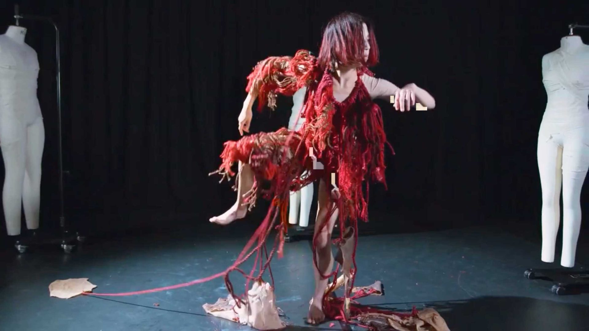 A person in a tattered, red costume poses dramatically onstage, surrounded by two white dress forms against a dark background. Fabric scraps are scattered on the floor, creating a striking scene inspired by The Midnight Club’s "Make a Life, Not Just a Living" for University of the Arts London. Frame from Stash Magazine article.