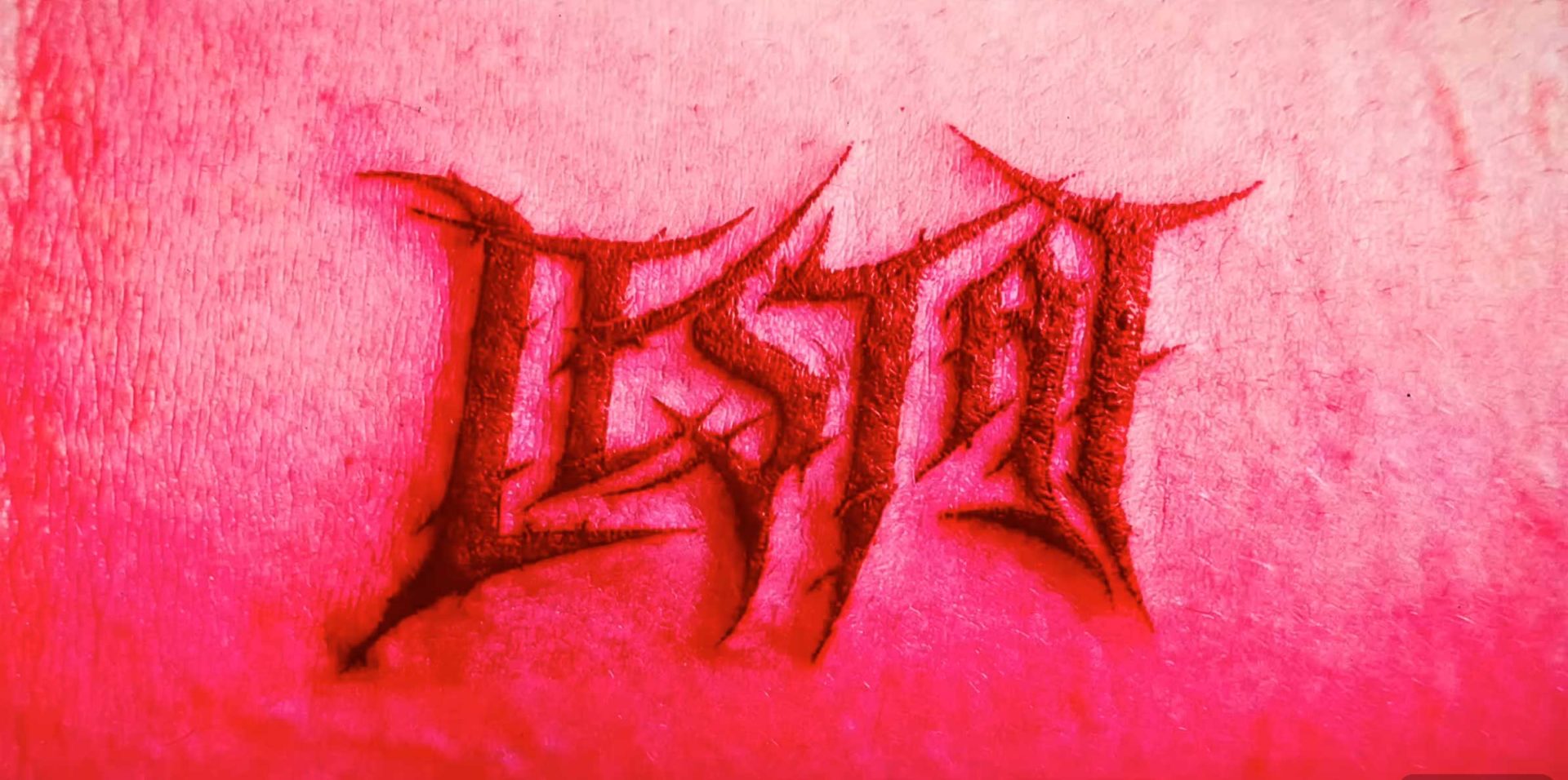 The word LESTAT is carved in jagged, stylized letters into pinkish skin, resembling a fresh scarification or tattoo—as if inspired by Anne Rice’s The Vampire Lestat—red tones highlighting the irritated area. Frame from Stash Magazine article.