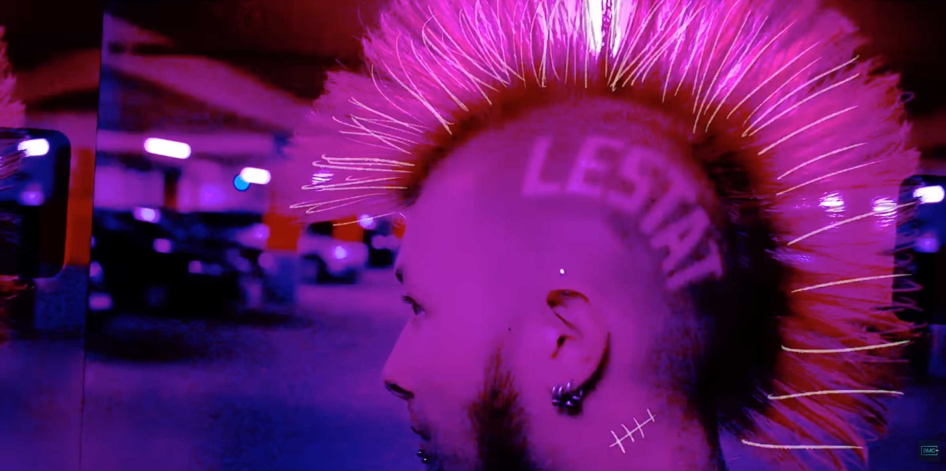 A person with a bright pink mohawk hairstyle and the word LESTAT—an homage to Anne Rice's The Vampire Lestat—shaved into the side of their head stands in a parking garage bathed in purple and pink lighting. Frame from Stash Magazine article.