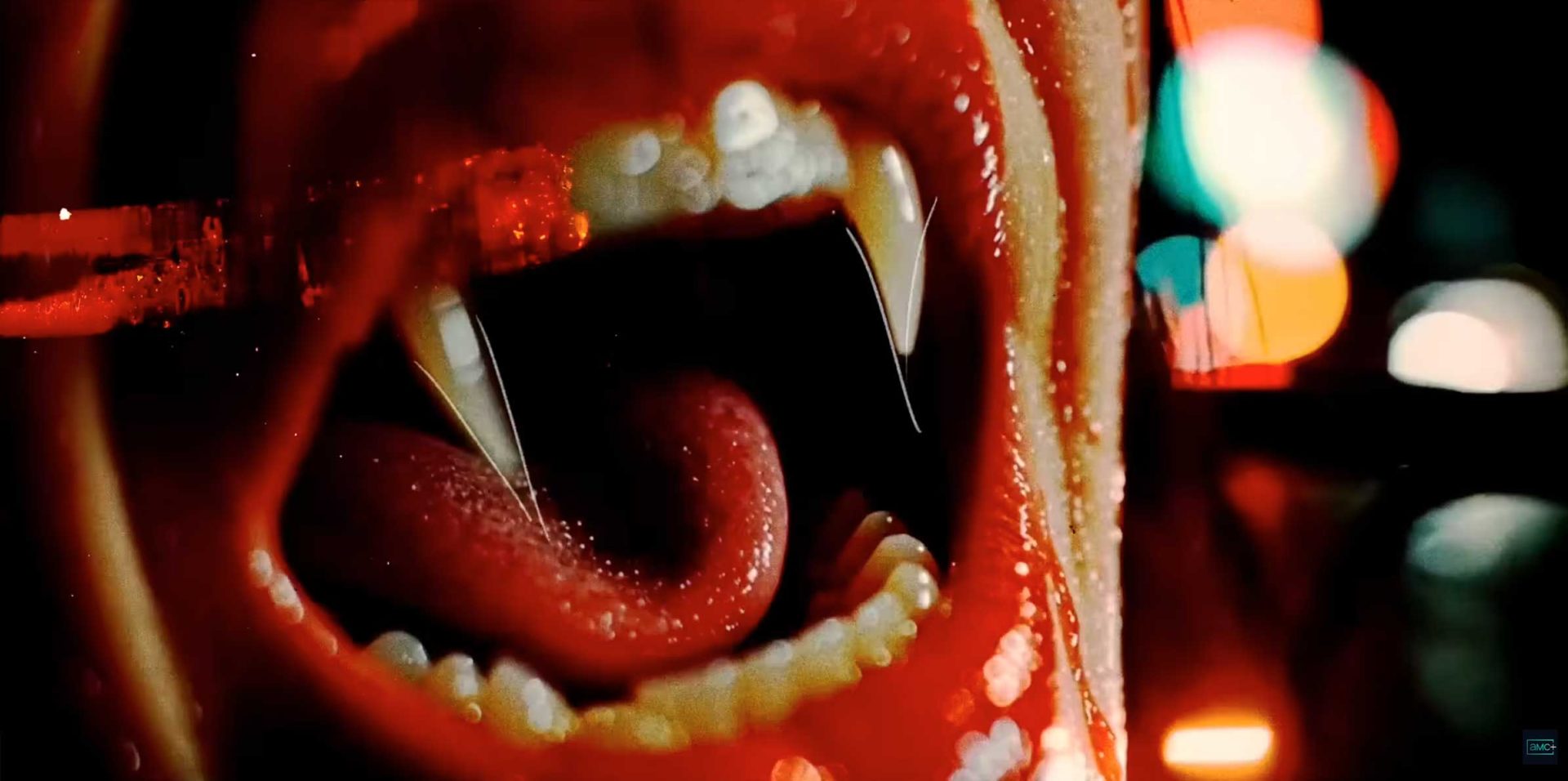 A close-up of an open mouth with sharp vampire fangs, a protruding tongue, and glossy red lips, reminiscent of The Vampire Lestat from Anne Rice's novels, with dramatic lighting and blurred colorful lights in the background. Frame from Stash Magazine article.