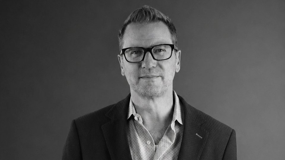 A man wearing glasses, a suit jacket, and an open-collared shirt stands in front of a plain dark background, looking directly at the camera with a neutral expression—capturing the professional poise often associated with Tobin_Kirk_EP_WORKPRODUCT-. Frame from Stash Magazine article.