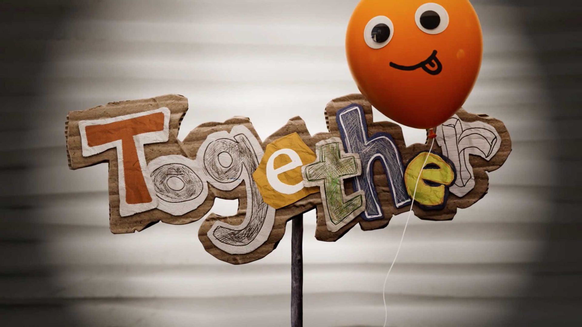 The word Together spelled out in cutout letters on a cardboard sign from the short film, topped with an orange balloon featuring a silly face. Frame from Stash Magazine article.