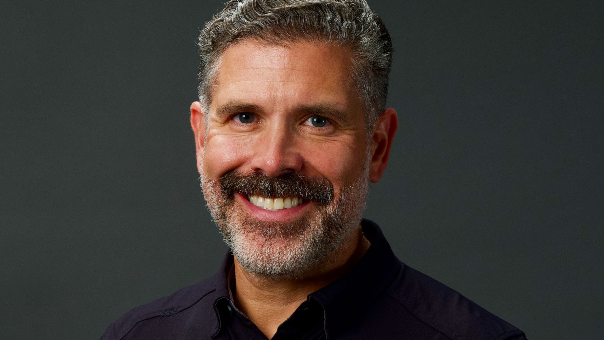 Headshot of Tony Kadillak, a smiling middle-aged man with short grey hair and a beard, set against a dark background. Frame from Stash Magazine article.