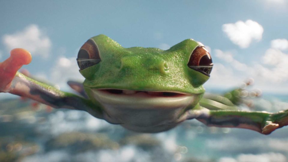 A close-up of a green tree frog with wide eyes and outstretched legs, looking like it leapt straight from a Tropicana Give Life Some Juice commercial by Dorian & Daniel and Untold, as it smiles toward the camera against a blurred outdoor background. Frame from Stash Magazine article.