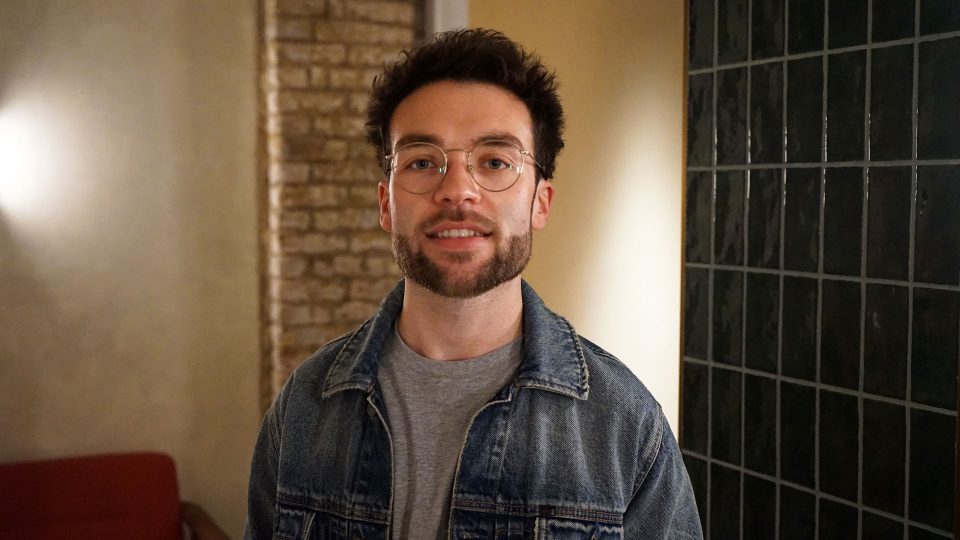 VFX Supervisor Alfie Vaughan joins RASCAL—a man with glasses, a beard, and short dark hair wears a denim jacket over a gray shirt, standing indoors by a tiled wall and exposed bricks. Frame from Stash Magazine article.