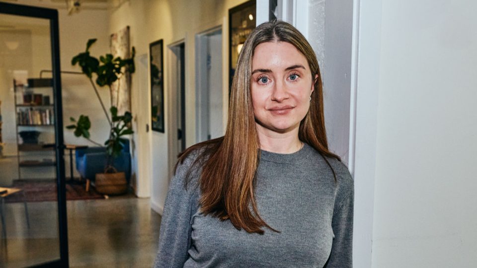 A woman with straight, long brown hair and a grey sweater stands indoors by a white wall, with modern office decor and plants in the background—this is Amanda Pellegrini, Head of Creative Operations at Vagrants. Frame from Stash Magazine article.