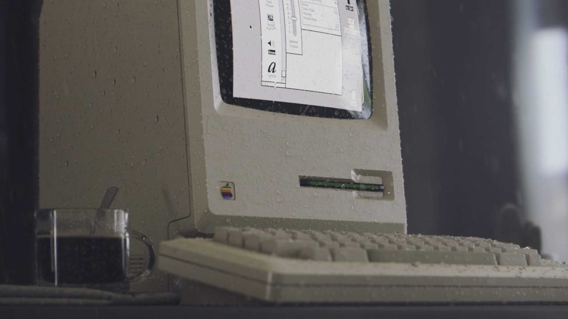 A vintage Apple Macintosh computer, part of Studio Aiko’s collection of vintage electronic devices, sits on a desk beside a glass cup of coffee. Water droplets cover the scene, and the screen shows an old graphical user interface. Frame from Stash Magazine article.