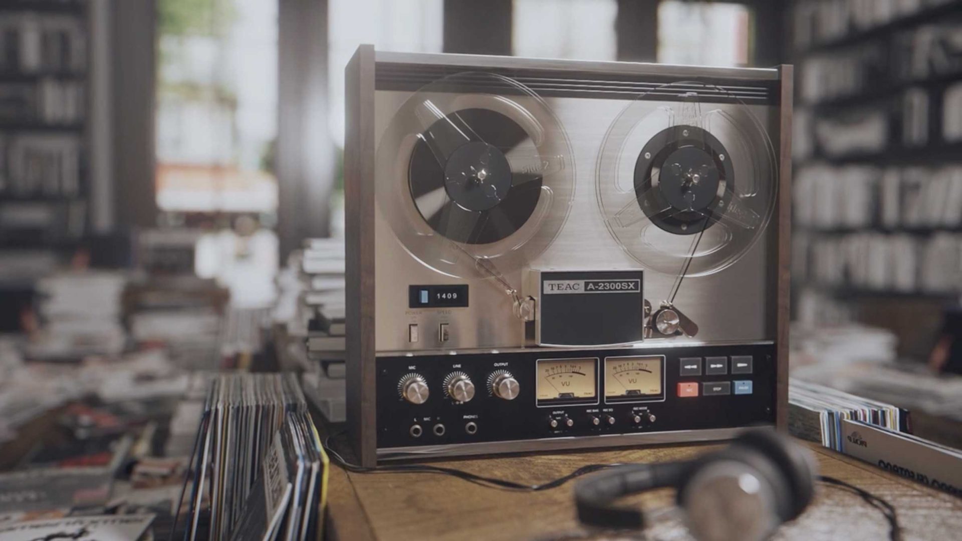 A vintage reel-to-reel tape recorder from Studio Aiko sits on a wooden table surrounded by stacks of books, vinyl records, and other vintage electronic devices, with headphones resting nearby in a cozy, sunlit room. Frame from Stash Magazine article.