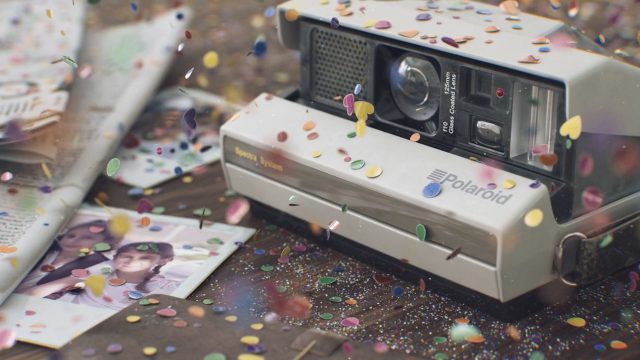 A vintage Polaroid camera sits on a wooden table covered with colorful confetti and sparkles, reminiscent of a scene from the short film "Vintage Electronic Devices" by Studio Aiko, alongside a developed photo showing two smiling people. Frame from Stash Magazine article.