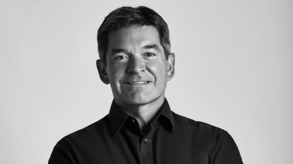 A man with short dark hair, wearing a dark button-up shirt, smiles slightly while looking at the camera. The image, in black and white, marks WPP Production's appointment of John Paulson as Managing Director in New York. Frame from Stash Magazine article.