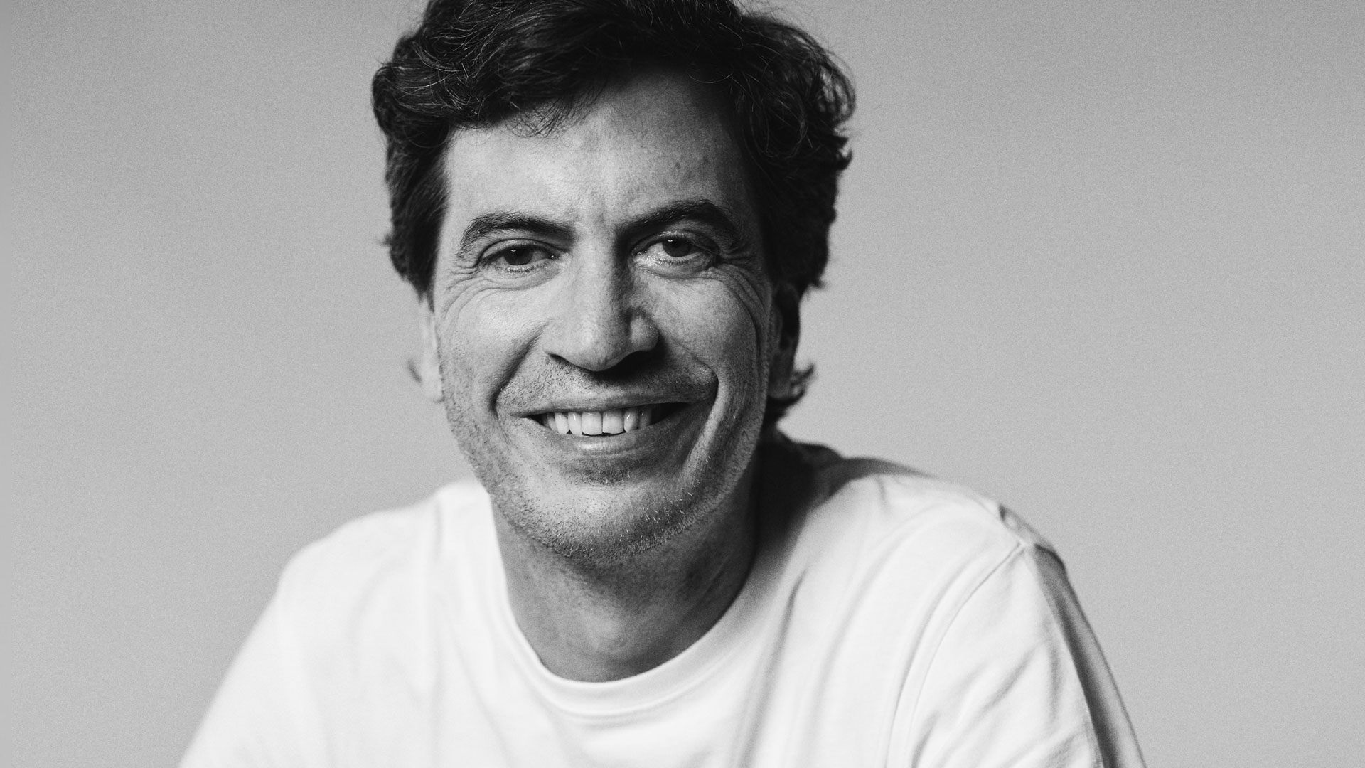 A man with wavy dark hair smiles warmly at the camera. He is wearing a plain white t-shirt and is photographed against a neutral, light background, capturing the approachable style of WPP Production's newly appointed CEO, Santiago Sánchez-Lozano for EMEA. Frame from Stash Magazine article.