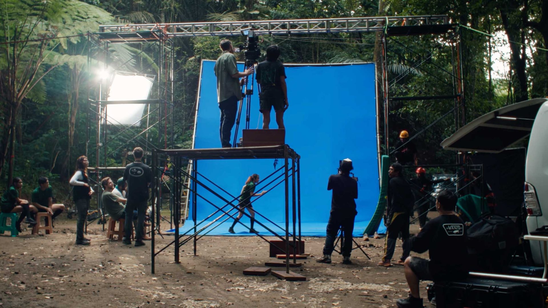 A film crew sets up a scene outdoors in a rainforest canopy, with a blue screen backdrop and lighting equipment. Two people stand on a metal platform with a camera, while others work around the set surrounded by trees and gear. Frame from Stash Magazine article.