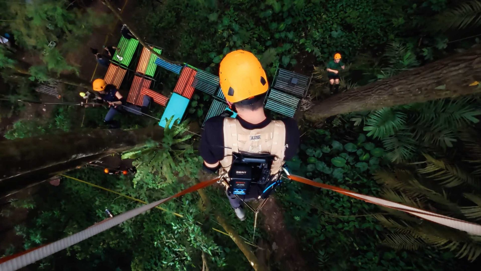 A person wearing a yellow helmet and safety harness stands on a high tree platform, preparing for a zip line adventure above the lush rainforest canopy—an area as vast as a football pitch. Colorful benches and three others are visible below in the trees. Frame from Stash Magazine article.
