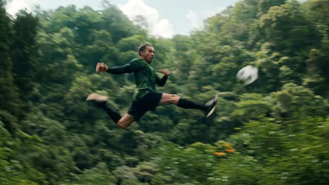 A soccer player in a green jersey and black shorts leaps horizontally in midair to kick a ball, set against the vibrant rainforest canopy, resembling a unique football pitch amidst nature. Frame from Stash Magazine article.