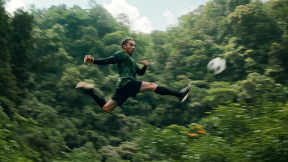 A soccer player in a green jersey and black shorts leaps horizontally in midair to kick a ball, set against the vibrant rainforest canopy, resembling a unique football pitch amidst nature. Frame from Stash Magazine article.