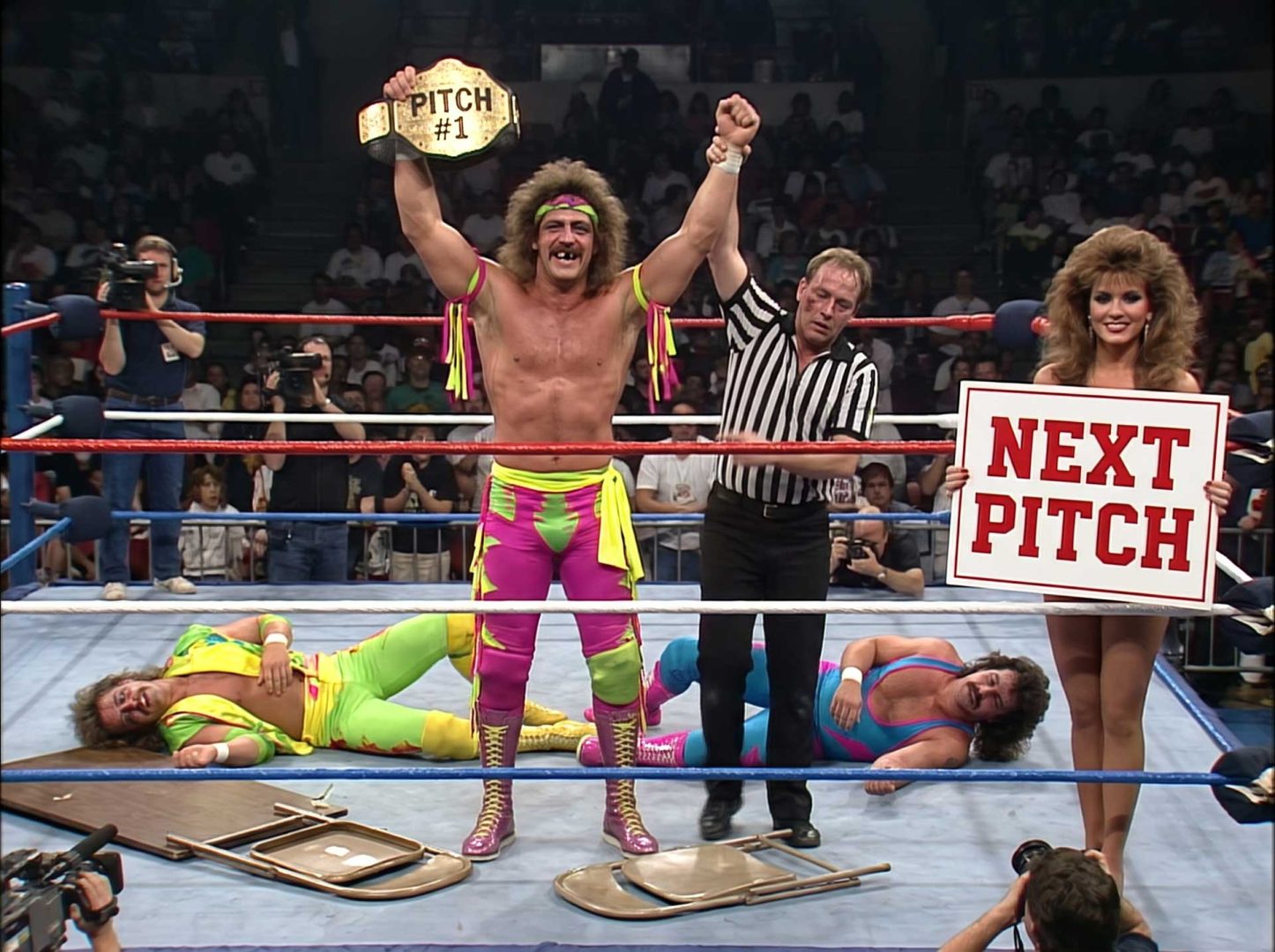 A wrestler in pink and yellow gear holds a championship belt labeled PITCH #1 in a ring with a referee, a woman with a NEXT PITCH sign, and two downed wrestlers, marking another scene from We-Love-Pitching-Part-23. Frame from Stash Magazine article.