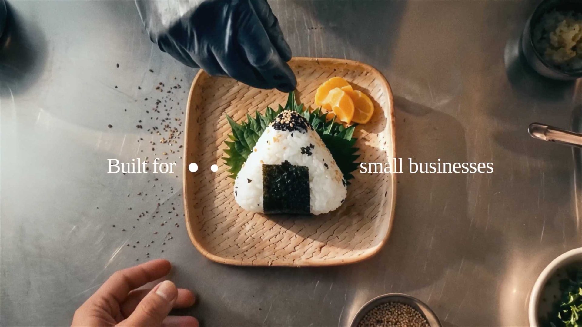 A gloved hand sprinkles seeds on a rice ball with seaweed on a square plate, garnished with orange slices and leaves; text reads Built for small businesses—brought to life with Wix Motion Studio. Frame from Stash Magazine article.