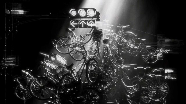 A dramatic black and white scene shows a chaotic pile of bicycles illuminated by beams of light, with traffic signals above them, creating a surreal and cluttered urban atmosphere reminiscent of New York’s restless energy. Frame from Stash Magazine article.