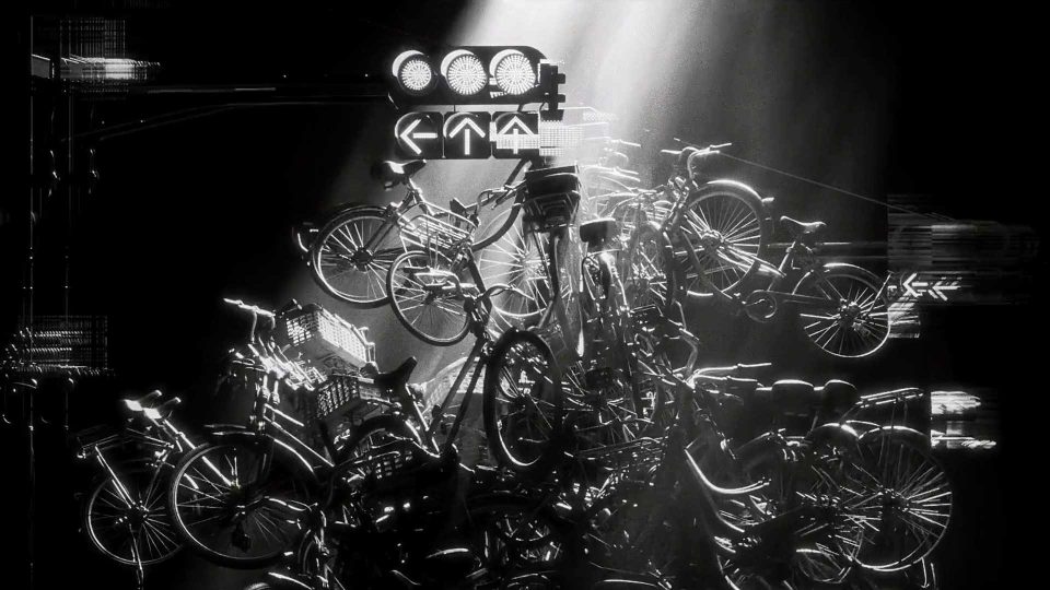 A dramatic black and white scene shows a chaotic pile of bicycles illuminated by beams of light, with traffic signals above them, creating a surreal and cluttered urban atmosphere reminiscent of New York’s restless energy. Frame from Stash Magazine article.